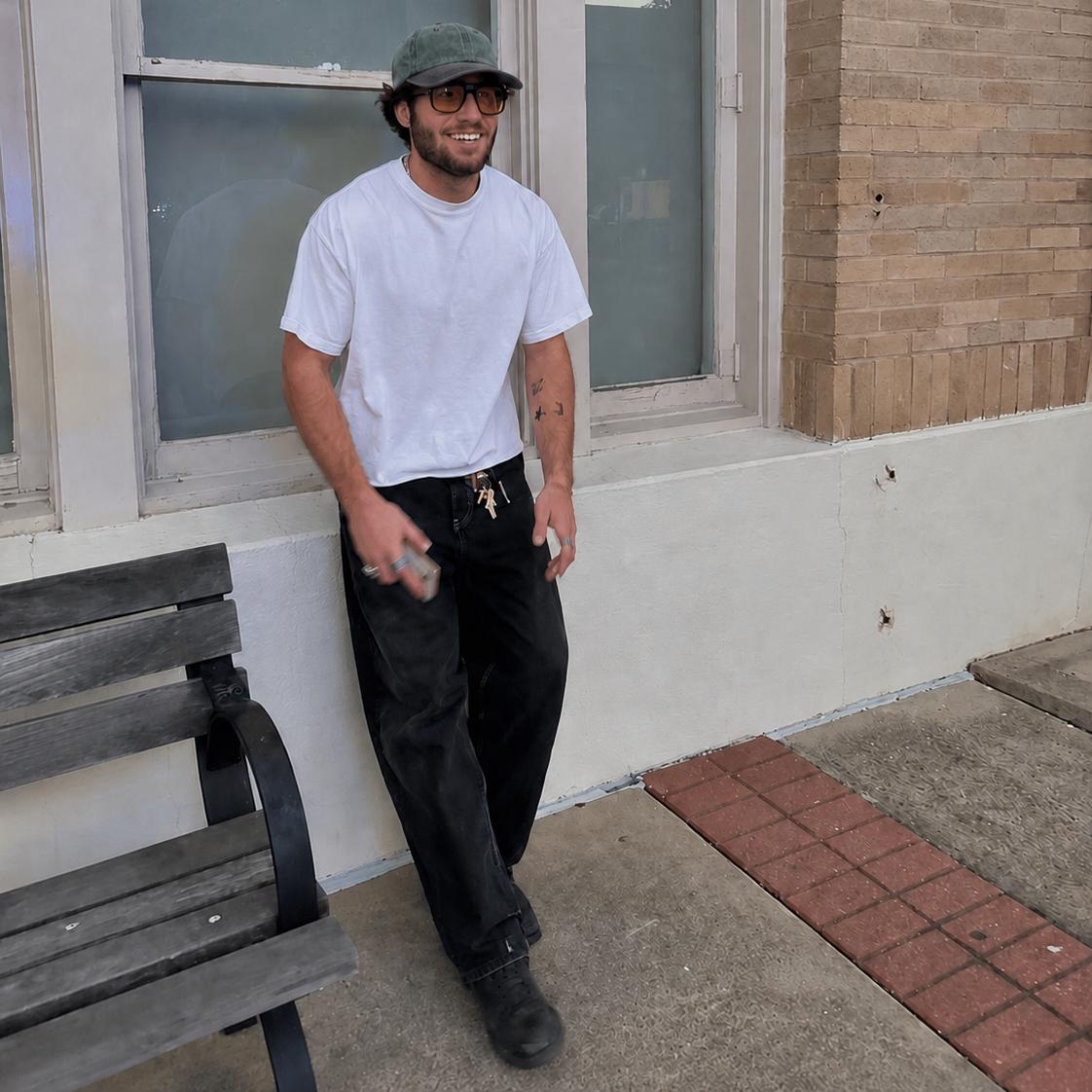 A man wearing a white crewneck t-shirt, black work pants, a green cap, and glasses stands on a sidewalk in front of a brick building.