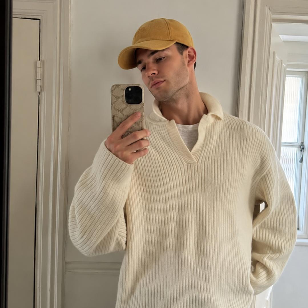 A man takes a mirror selfie wearing a cream ribbed knit polo sweater, olive green pants, and a mustard yellow baseball cap in a home setting.