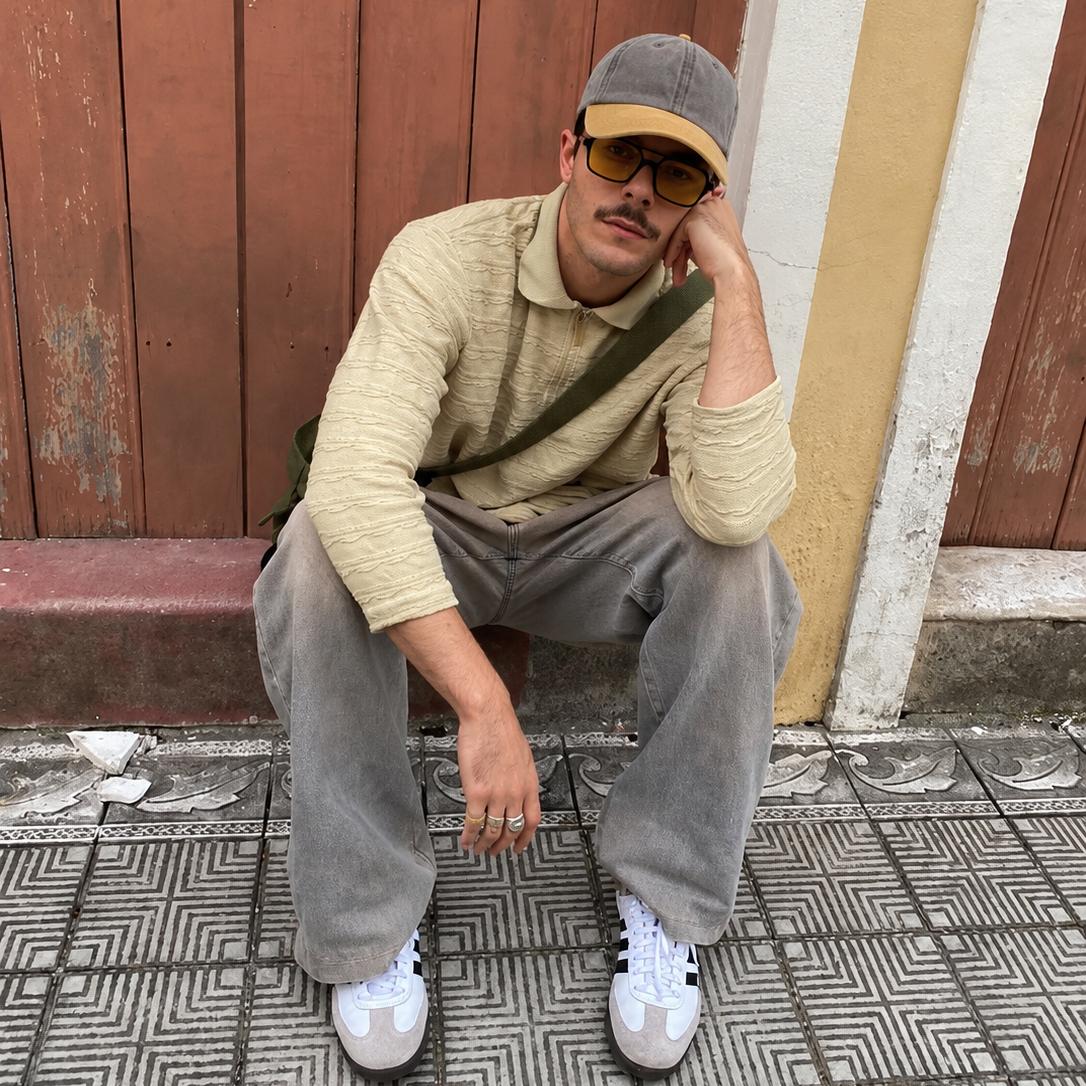 A man wearing a beige ribbed knit polo shirt, gray corduroy pants, white sneakers, and a gray cap with a yellow brim sits on a tiled ledge against a rustic wooden wall.
