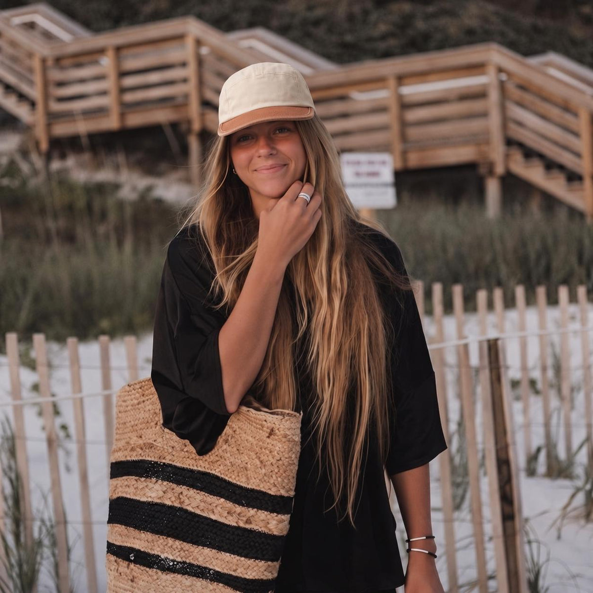A woman standing on a sandy beach wearing a cream baseball cap, a black short-sleeve top, and carrying a woven straw tote bag with black stripes. Wooden dune stairs and beach grass are visible in the background.