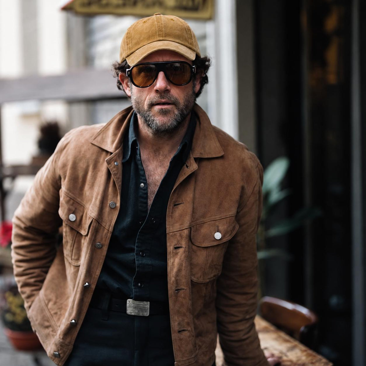 A man wearing a brown suede jacket, black shirt, black pants, sunglasses, and a tan baseball cap, standing in an outdoor urban setting.