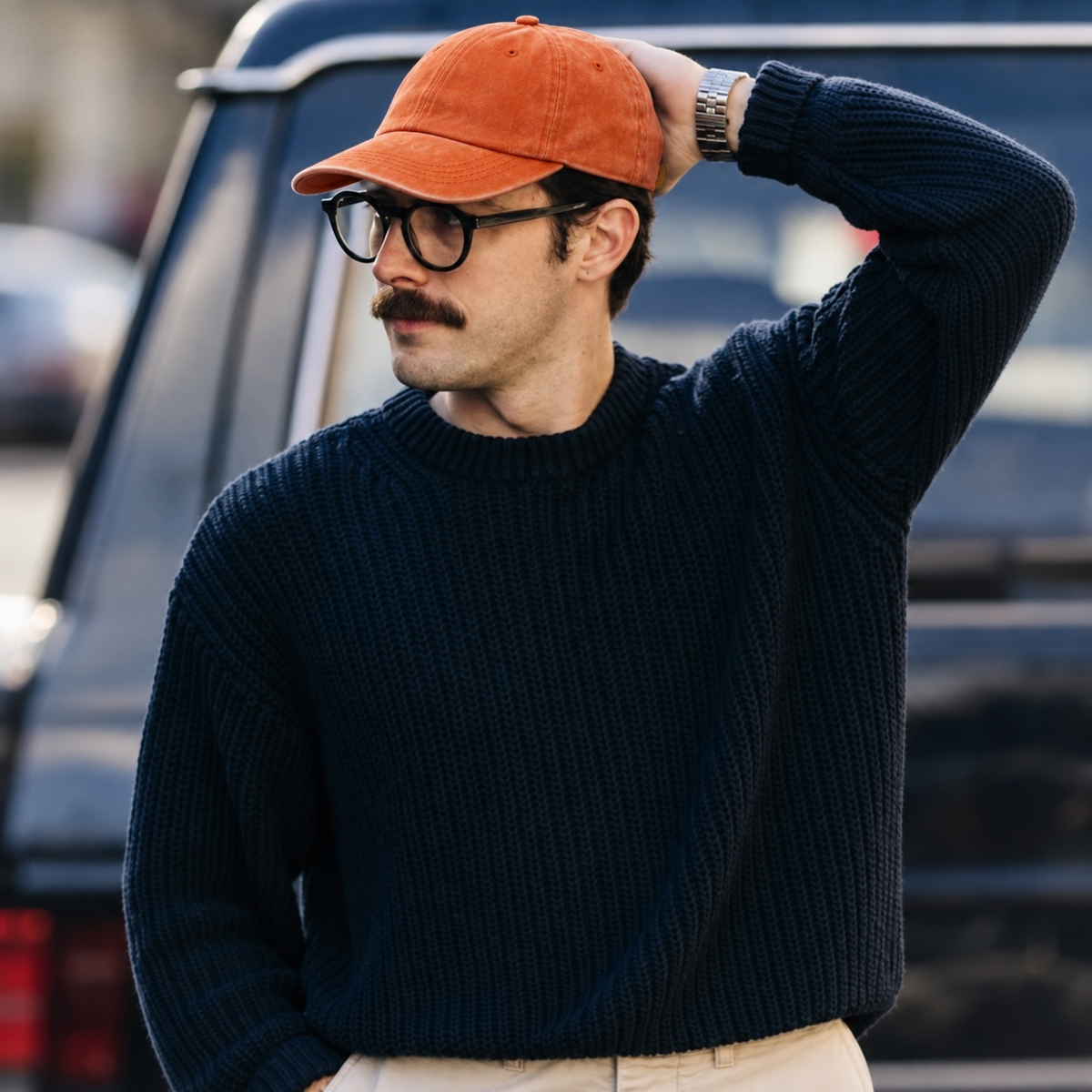 A man wearing a dark navy ribbed knit sweater, an orange baseball cap, and black-rimmed glasses, standing outdoors in front of a dark vehicle.