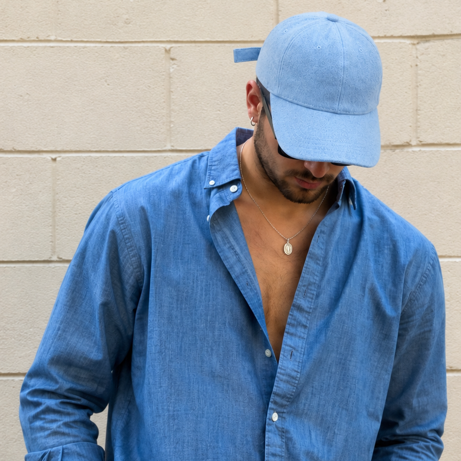 A man wearing a light blue chambray button-down shirt, dark navy athletic shorts, and a matching light blue baseball cap, standing against a beige brick wall.
