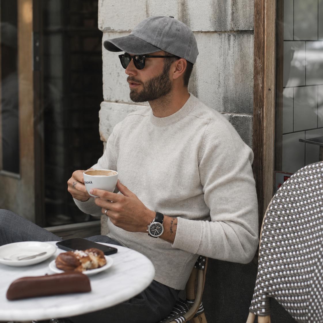 A man sitting at an outdoor cafe table wearing a light gray baseball cap, sunglasses, a beige knit sweater, and a watch, holding a white coffee cup.