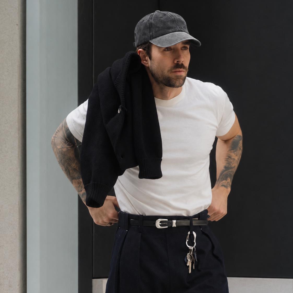 A man wearing a white crew-neck t-shirt, dark navy trousers, a black belt, and a gray baseball cap, with a black jacket draped over his shoulder, standing against a dark modern wall.