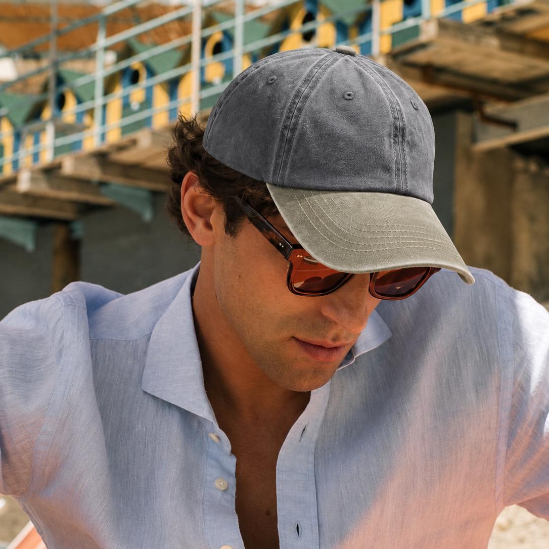 A man wearing a gray and beige baseball cap, sunglasses, and a light blue linen shirt, photographed in a real outdoor setting with stadium seating in the background.