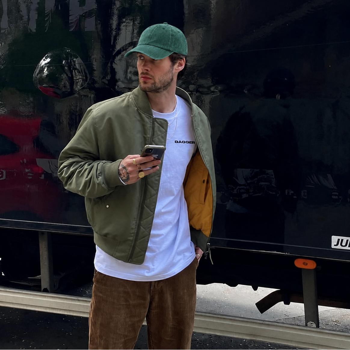 A man wearing a green baseball cap, olive green bomber jacket, white t-shirt, and brown corduroy pants stands in front of a black truck, holding a smartphone.