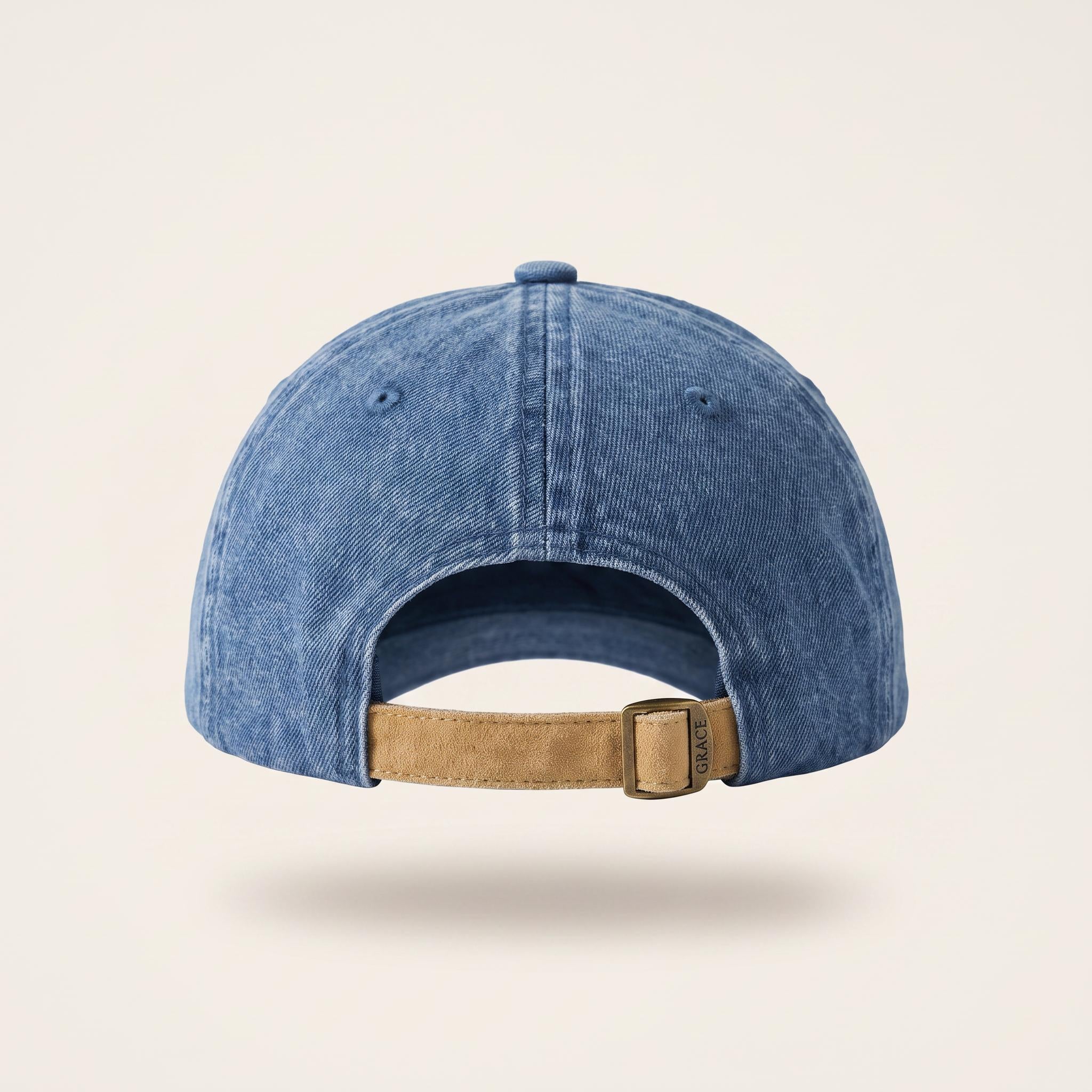 A blue washed denim baseball cap shown from the back, featuring a tan leather strap and a silver metal buckle, set against a light beige background.