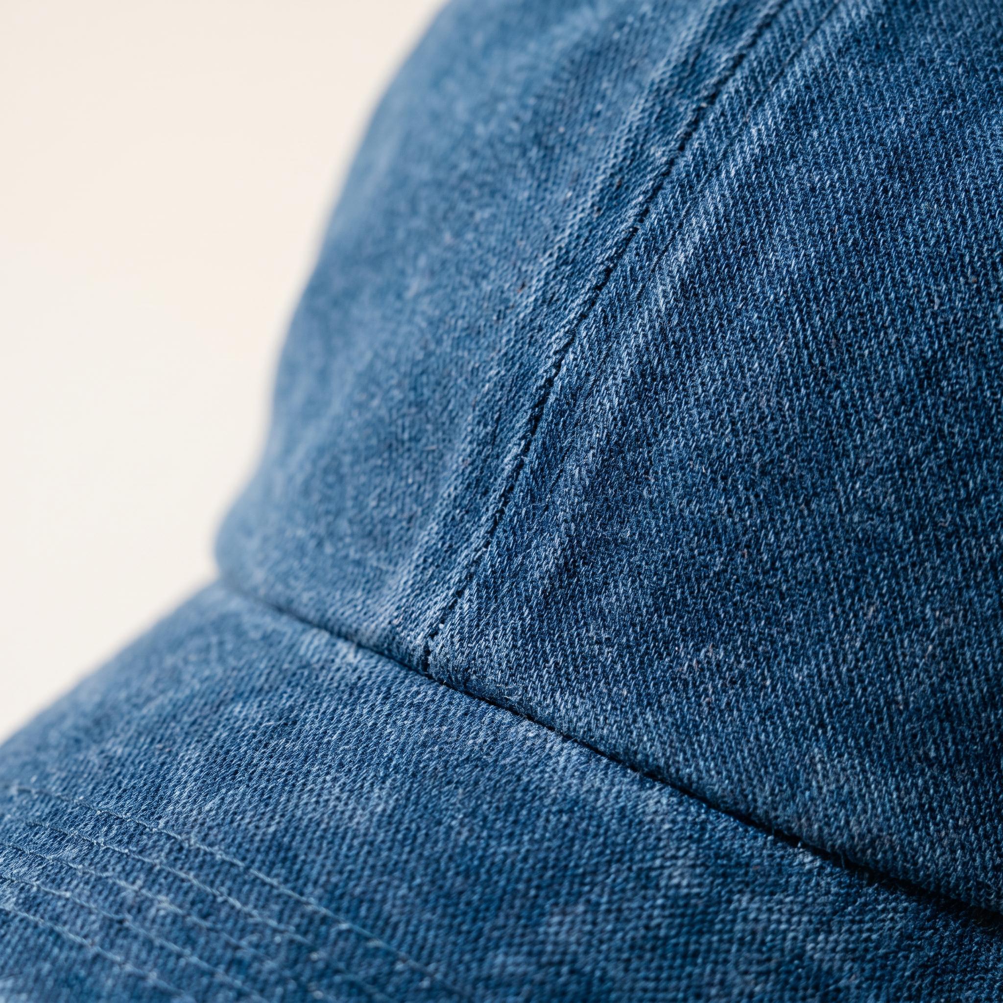 Close-up detail of a blue denim baseball cap showing the fabric texture, curved brim, and stitched panel seams against a light cream background.