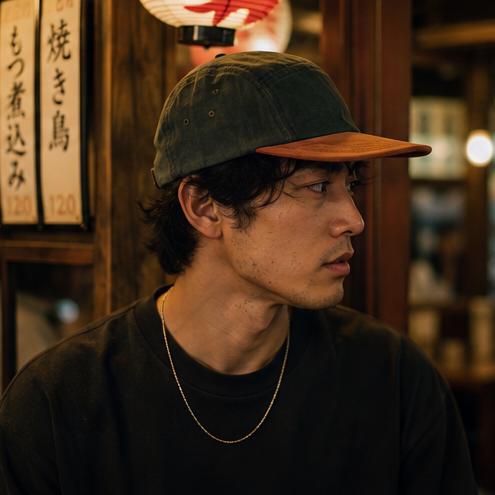 A man in a dark green and brown baseball cap, wearing a black crewneck sweatshirt and a gold chain necklace, photographed in a warm, dimly lit Japanese restaurant setting.