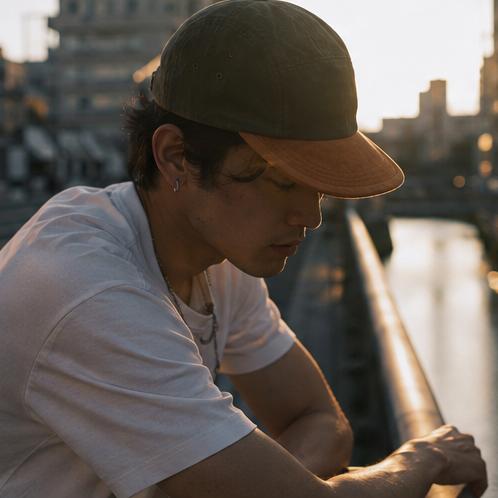 A man wearing a dark olive green baseball cap and a white t-shirt leans over a railing next to a canal during golden hour, with a blurred cityscape in the background.
