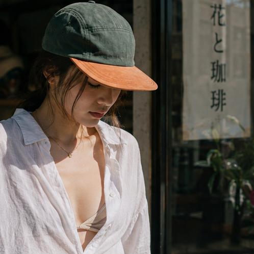 A woman wearing a dark green baseball cap with a contrasting orange brim, an open white button-up shirt, and a beige bikini top, standing in front of a shop window with Japanese text.