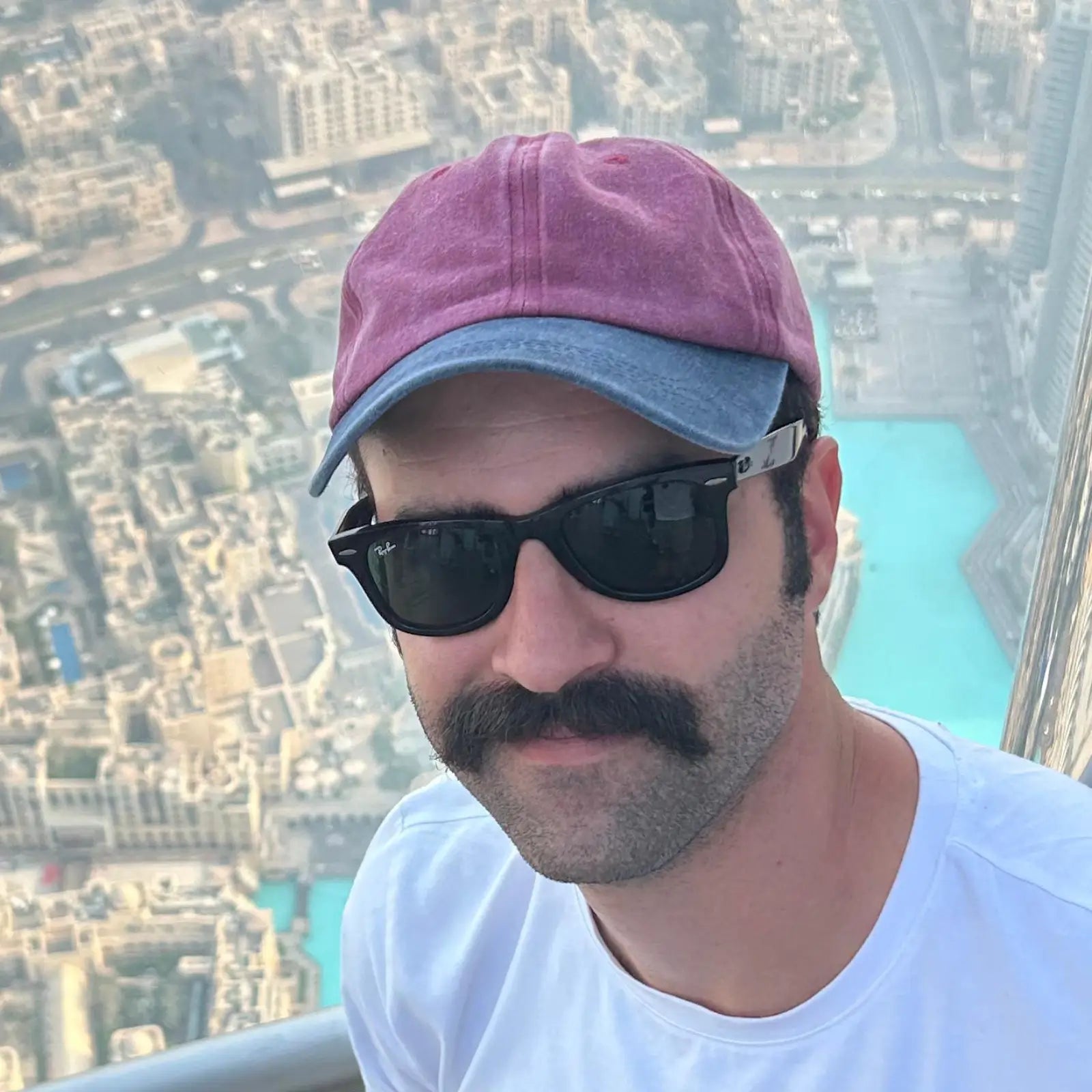 A man with a mustache wearing a dusty pink and blue baseball cap, black sunglasses, and a white t-shirt, photographed from a high-rise balcony overlooking a city skyline and turquoise water.