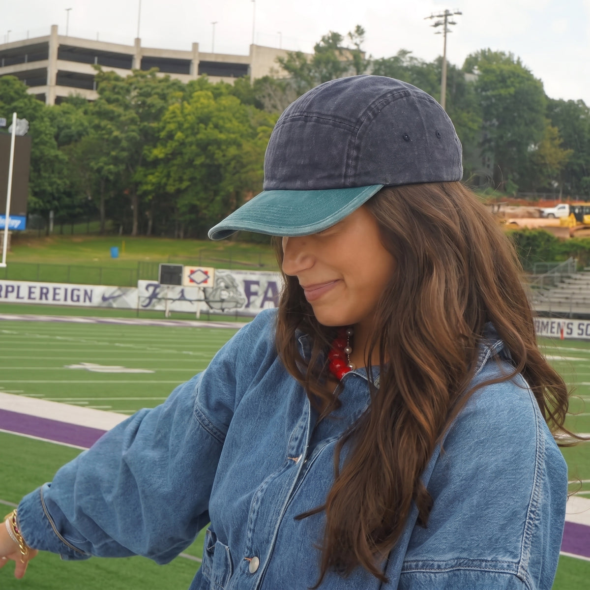 A woman wearing a washed navy blue baseball cap with a teal visor and a blue denim jacket, standing on a football field with trees and a parking structure in the background.