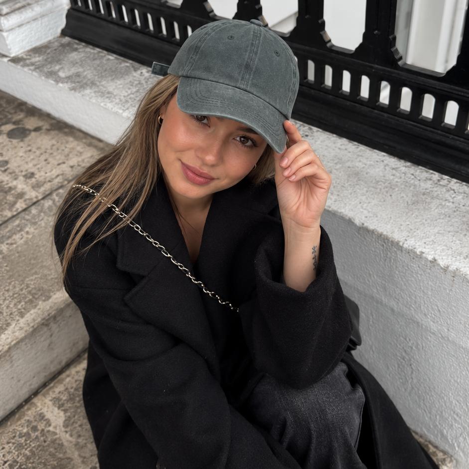 A woman wearing a dark gray baseball cap, black coat, and dark skirt sits on stone steps in front of a black metal railing.