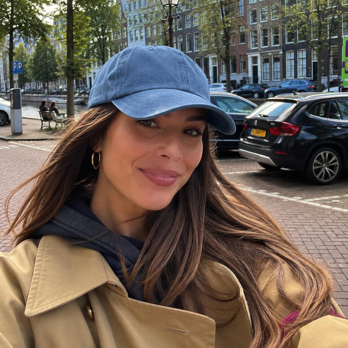 A woman wearing a light blue denim baseball cap, a tan trench coat, and a dark blue scarf, posing outdoors on a brick street with parked cars and buildings in the background.
