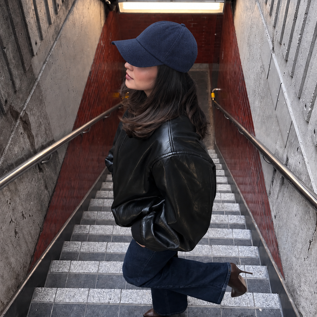 A woman wearing a navy blue baseball cap, black leather jacket, and dark blue jeans walks down a tiled staircase in a subway station, with a sign reading 'QUEEN' above her.