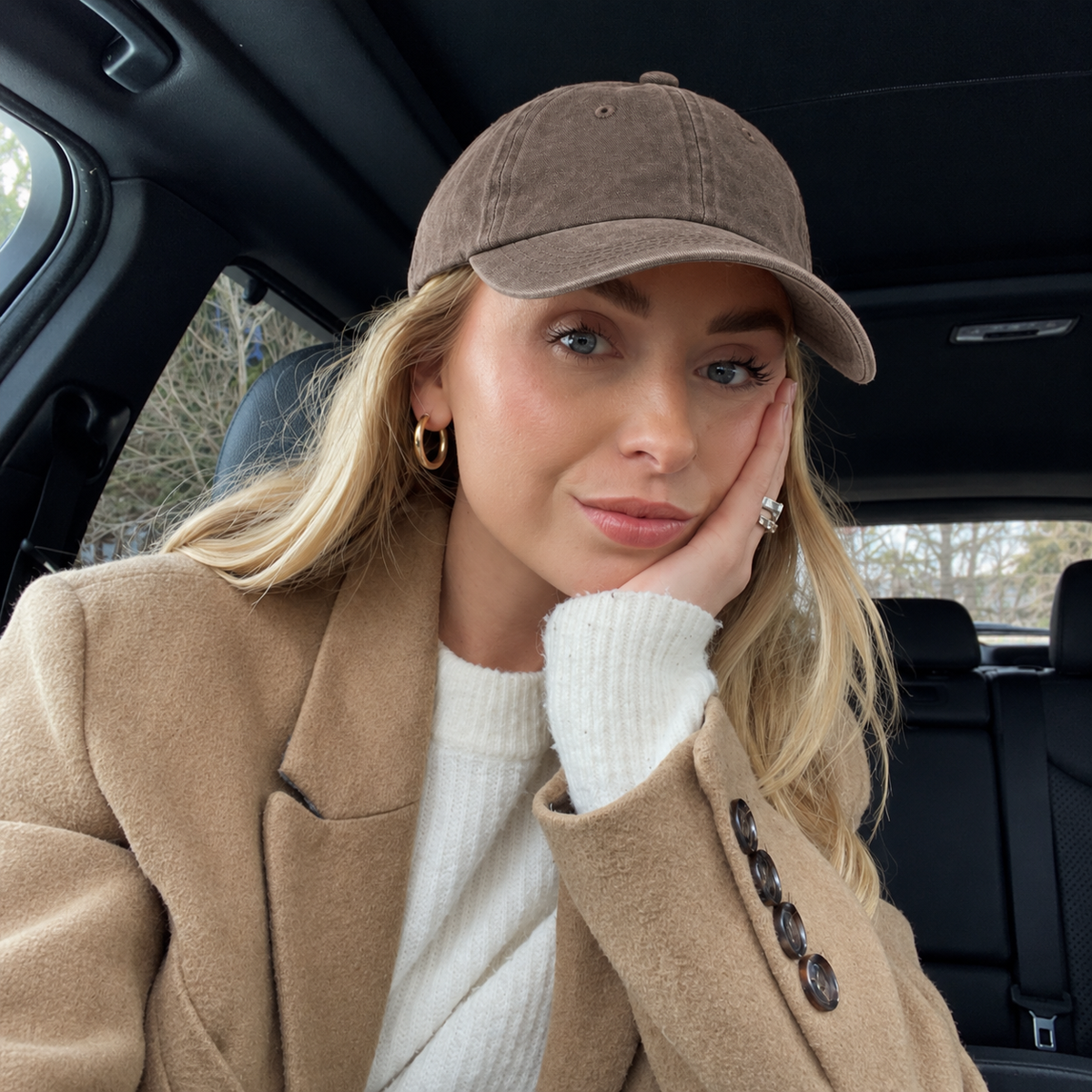 A woman sitting inside a car wearing a brown baseball cap, a camel-colored coat, and a white ribbed sweater.