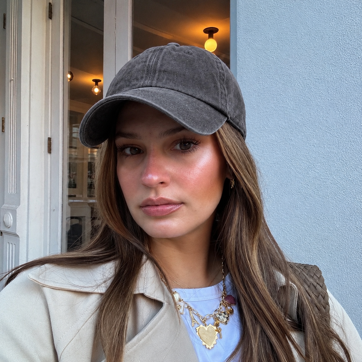 A woman wearing a washed dark gray baseball cap, a beige trench coat, and a white t-shirt with gold necklaces, photographed in an outdoor setting.
