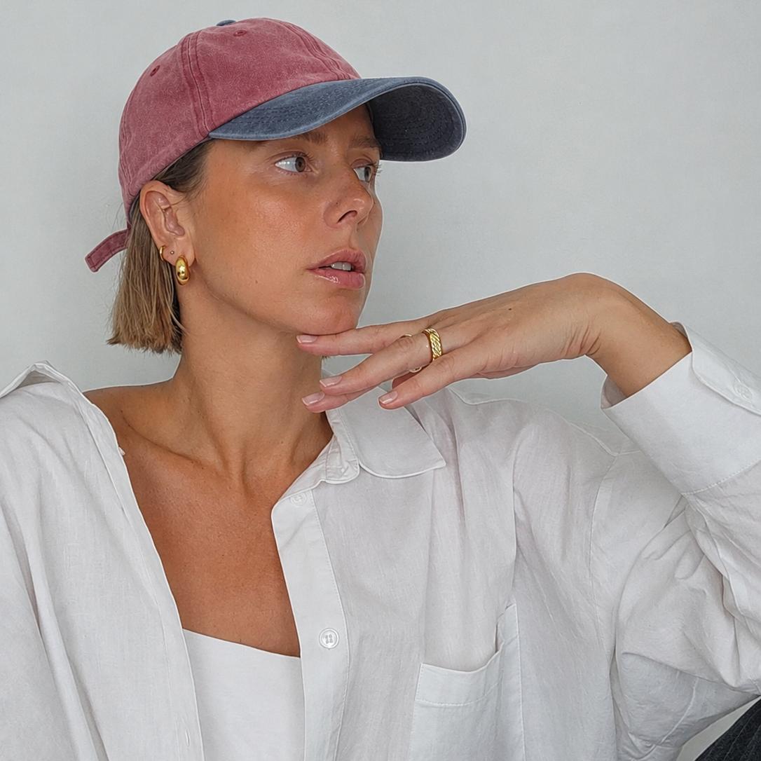 A woman wearing a dusty rose and navy blue two-tone baseball cap, a white button-up shirt, and a white tube top, posing against a light gray wall.