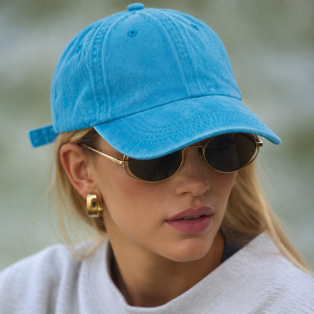 A close-up of a woman wearing a bright blue cotton baseball cap, gold-framed sunglasses, and a light gray sweatshirt.