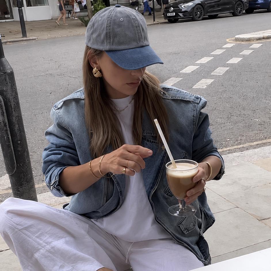 A woman sitting at an outdoor cafe table wearing a light blue denim jacket, white wide-leg pants, a blue cap, and blue slide sandals, holding a drink with a straw.