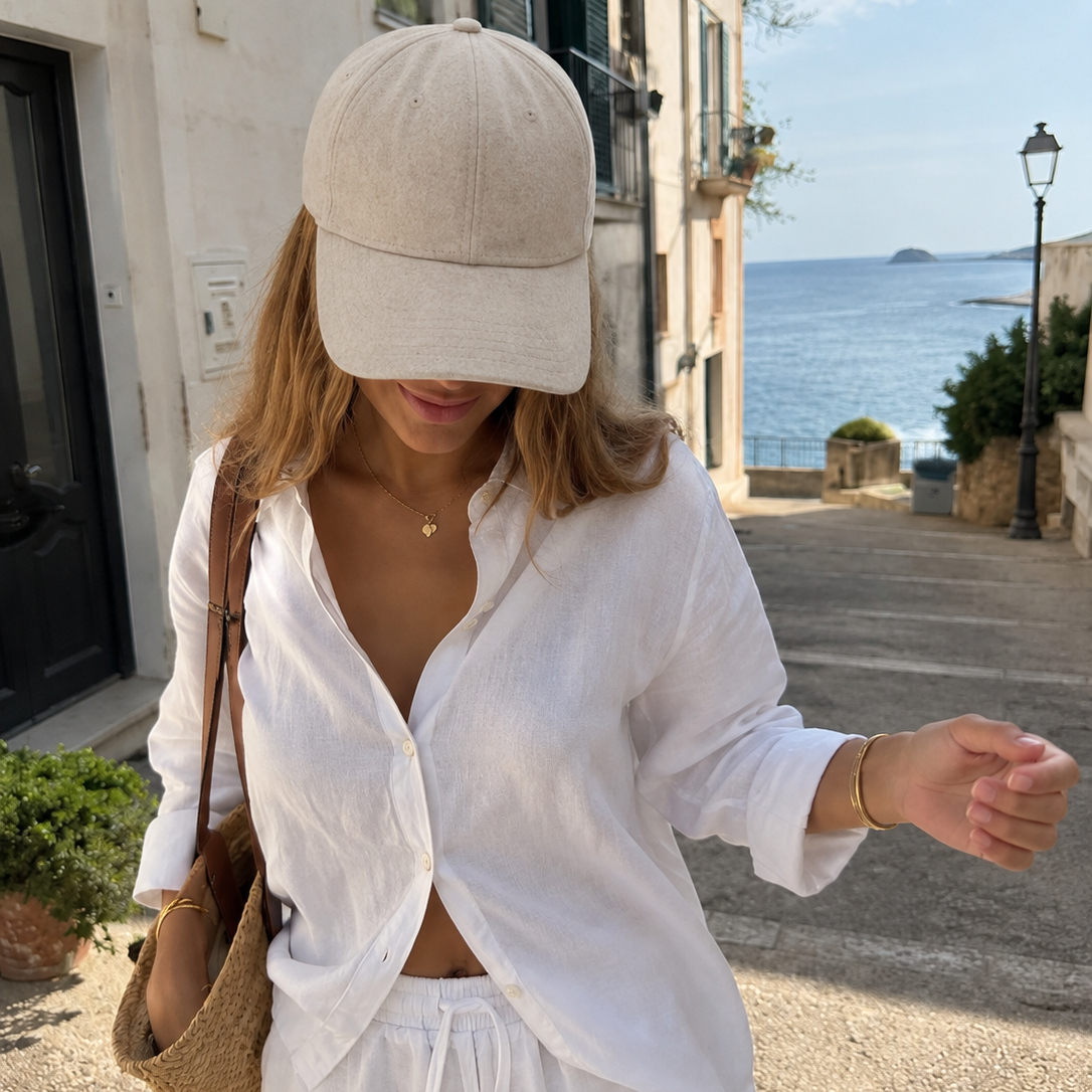 A woman walking outdoors on a sunny coastal street wearing a light beige baseball cap, a white button-up shirt, and white shorts, with a tan shoulder bag.