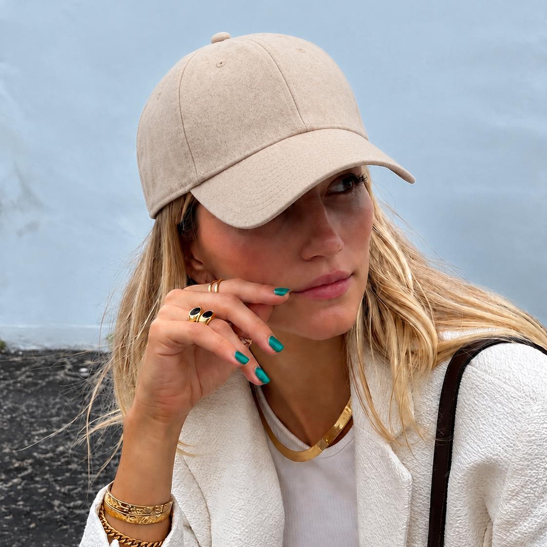 A woman wearing a beige baseball cap, a white textured jacket, and gold jewelry, posing outdoors against a soft blue-gray background.