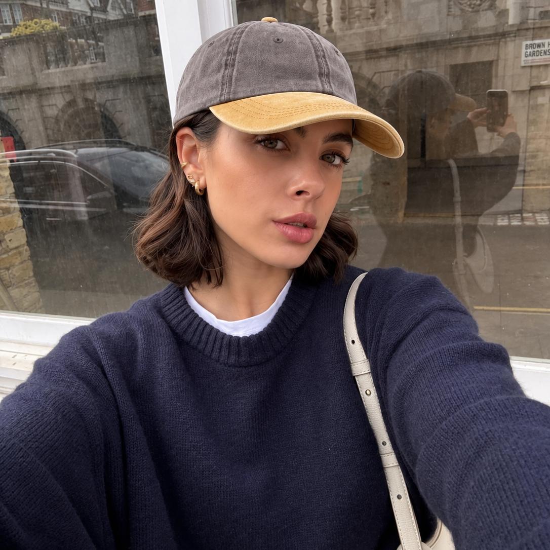 A woman wearing a gray and tan baseball cap, a navy blue knit sweater, and a white shoulder bag, taking a selfie in front of a window with a blurred street scene in the background.