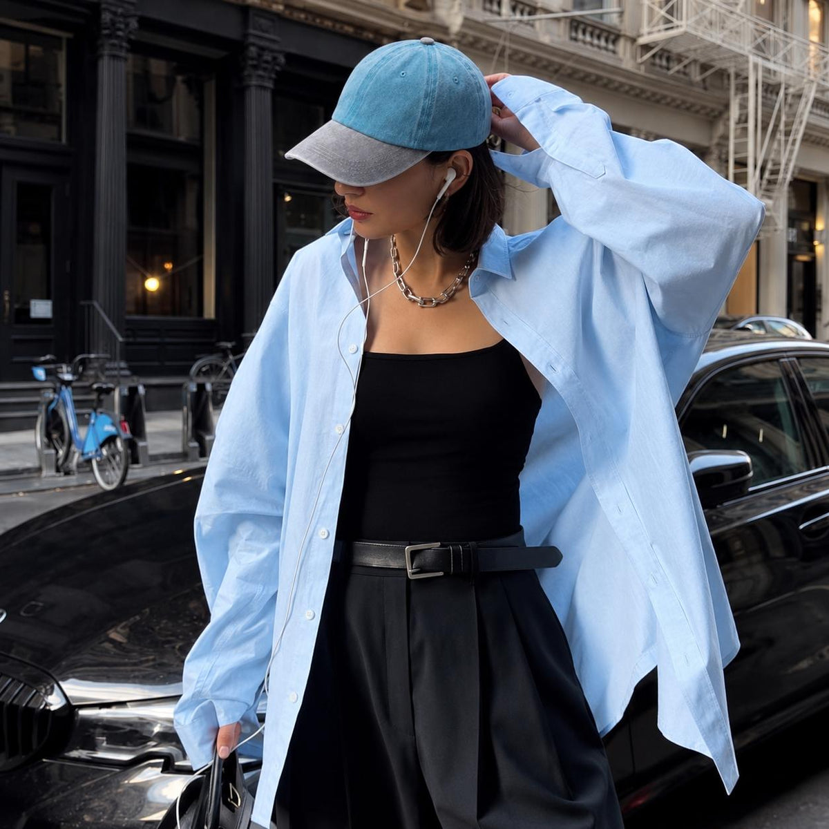 A woman standing on a city street wearing an oversized light blue button-up shirt over a black tank top and black skirt, with a blue and gray baseball cap, earphones, and a black belt.