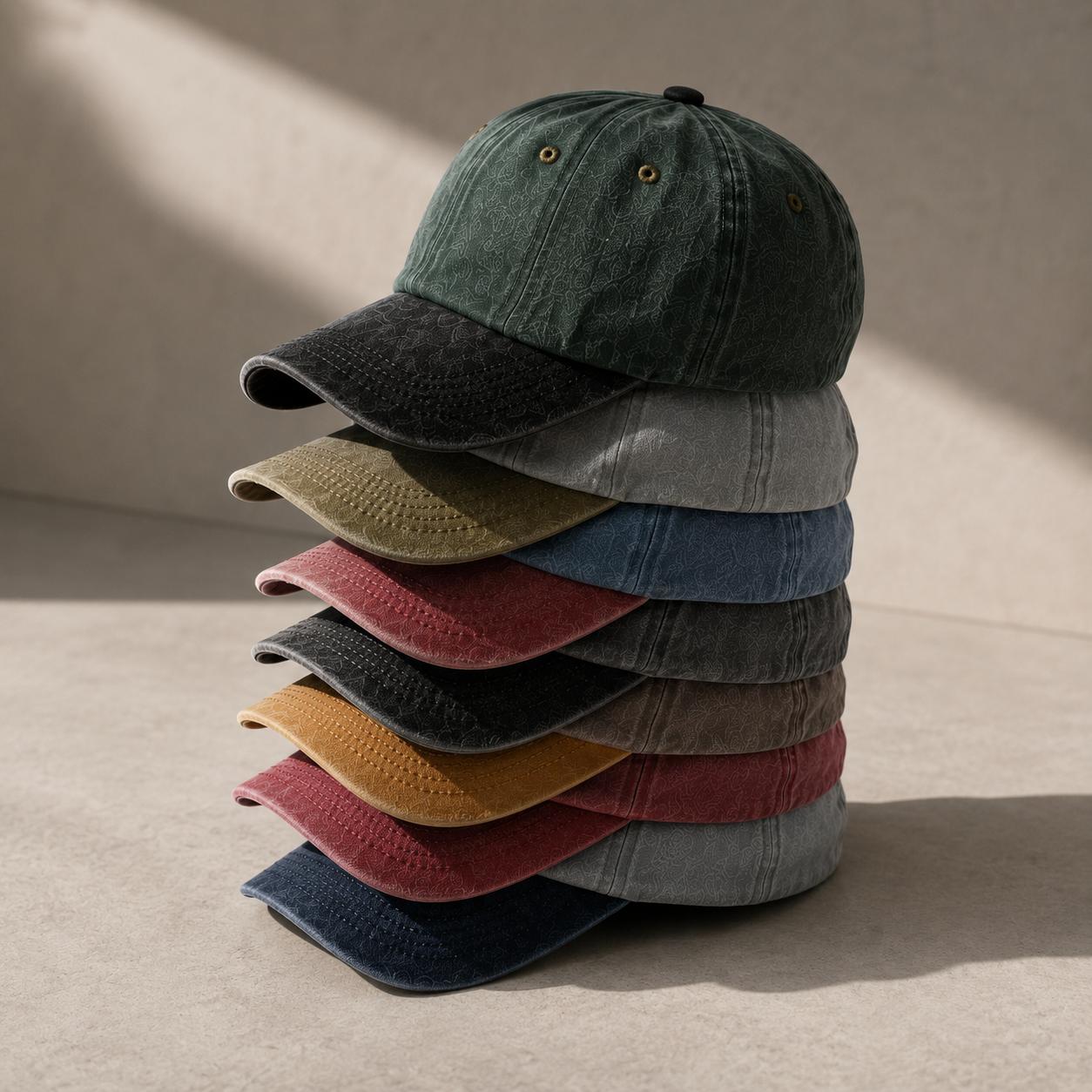 A stack of six baseball caps in different muted colors, including dark green, charcoal, olive, light gray, blue, burgundy, black, mustard, brown, and navy, arranged on a textured beige surface with soft diagonal shadows.