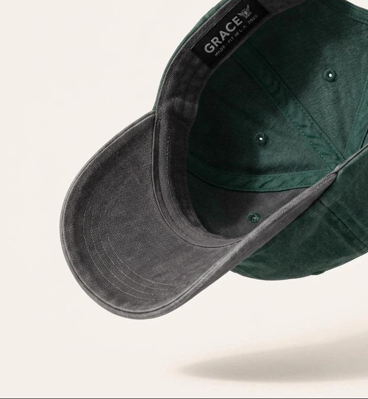 A dark forest green baseball cap with a washed, textured finish, shown floating at an angle to display both the curved brim and the interior. The brand name 'GRACE' is visible on a black label sewn inside the crown.