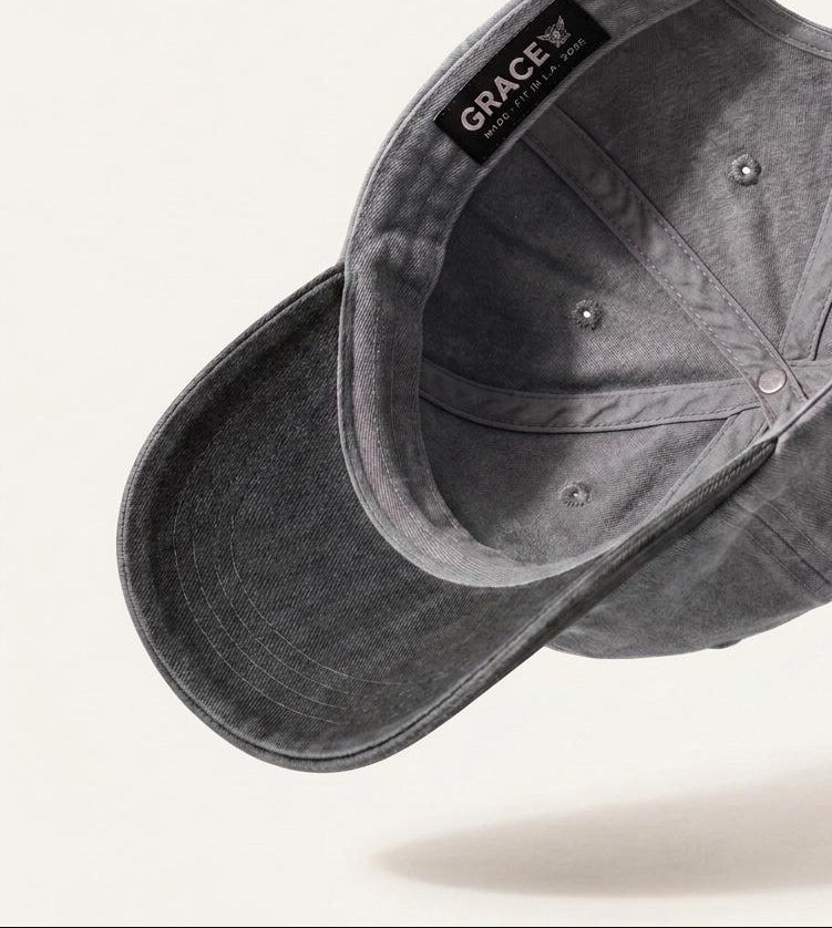 A dark gray washed cotton baseball cap shown floating at an angle, displaying the interior crown, snap closure, and a black interior label that reads 'GRACE'.
