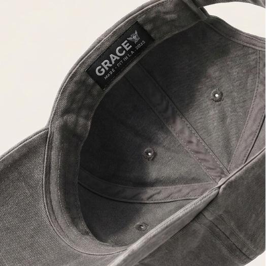 A dark gray washed cotton baseball cap floating at an angle against a light beige background, showing the interior with a black 'GRACE' brand label.