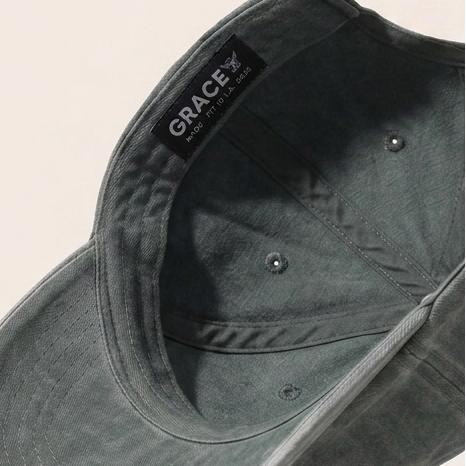 A dark green washed cotton baseball cap floating at an angle against a warm off-white background, with a black interior label reading 'GRACE'.
