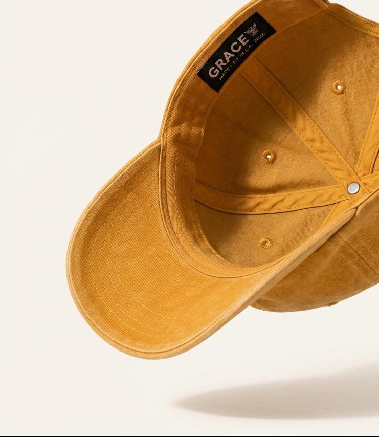 A mustard yellow baseball cap shown from an angled top-down perspective, floating against a clean off-white background. The brand name 'GRACE' is clearly visible on a black label stitched inside the crown.
