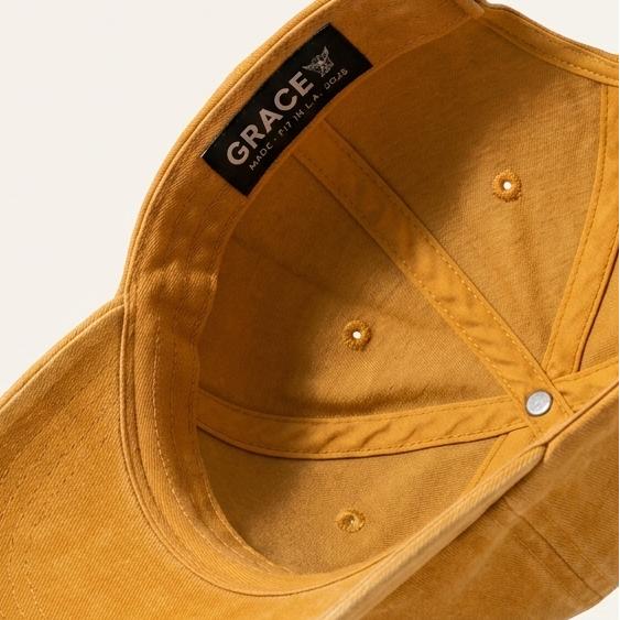 A mustard yellow baseball cap shown from an angled top-down perspective, floating against a clean off-white background. The brand name 'GRACE' is clearly visible on a black label stitched inside the crown.