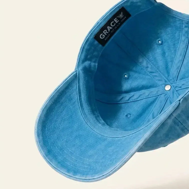 A light blue denim baseball cap shown from an angled top-down perspective, floating against a solid cream background. The brand name 'GRACE' is visible on a black interior label.