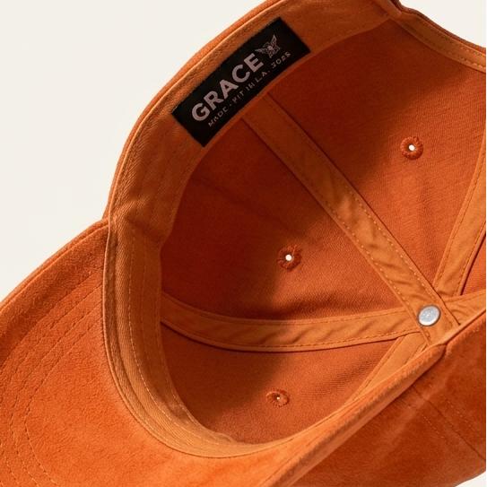 An orange suede baseball cap shown from an angled top-down view, floating against a light beige background with a soft shadow underneath. The brand name 'GRACE' is visible on a black label stitched inside the crown.