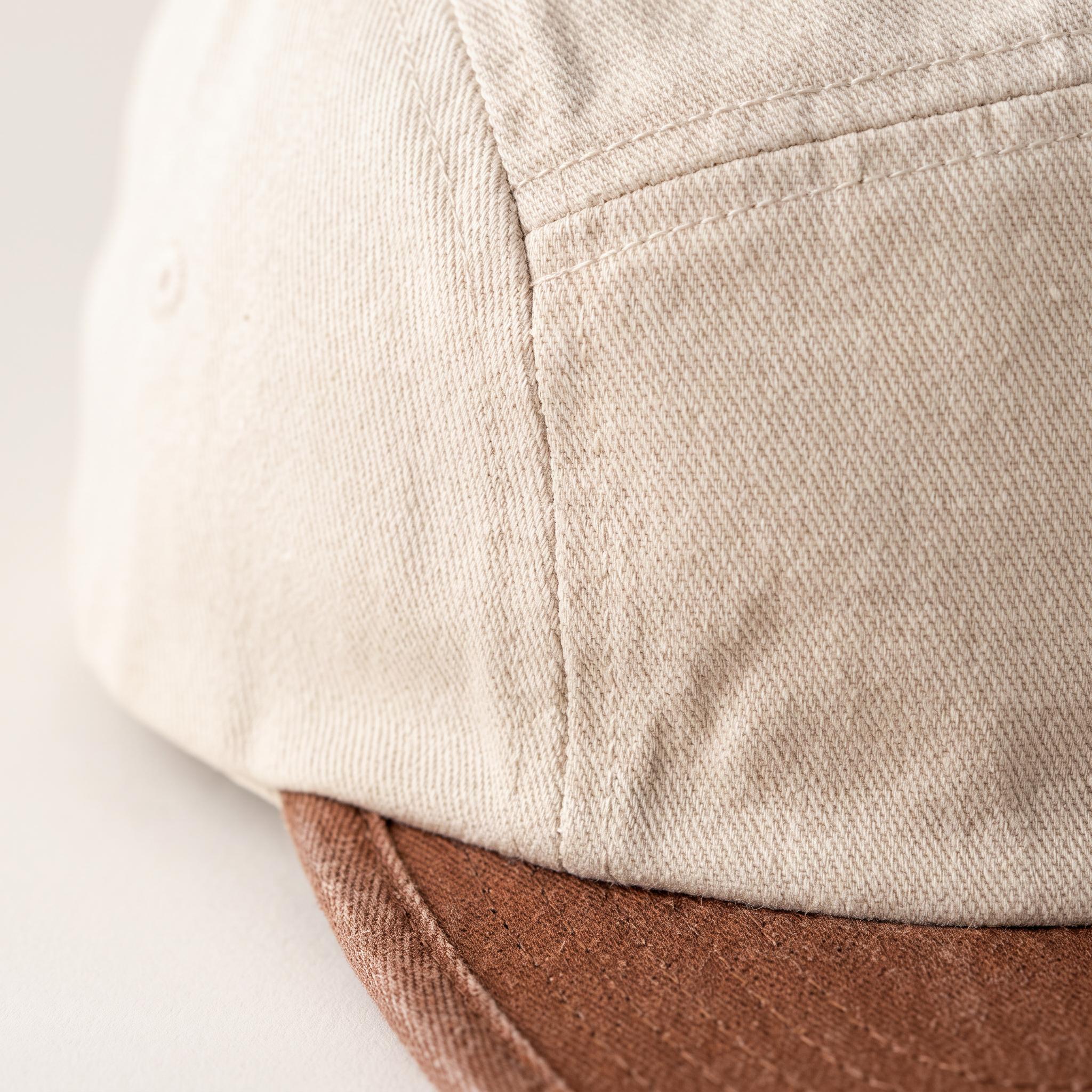 Close-up of a baseball cap showing the side panel in a light beige twill fabric and the edge of a brown fabric brim.
