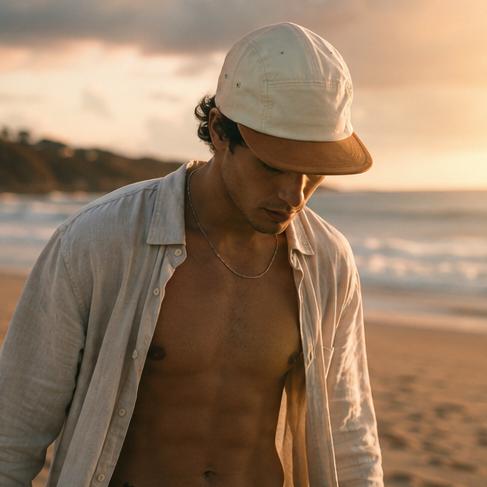 A man standing on a sandy beach at sunset wearing a light-colored baseball cap with a brown brim, an open light shirt, and a necklace.