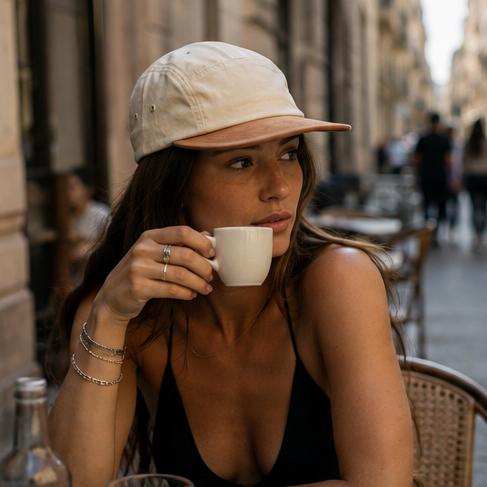 A woman sitting at an outdoor cafe table wearing a light beige baseball cap and a black top, holding a white coffee cup. The background shows a blurred street scene with cafe tables and people.