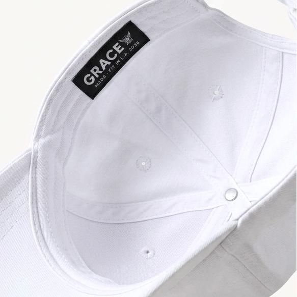 A white structured baseball cap shown floating at an angle against a light cream background, with a black interior label reading GRACE and a small gold metal accent near the back strap.