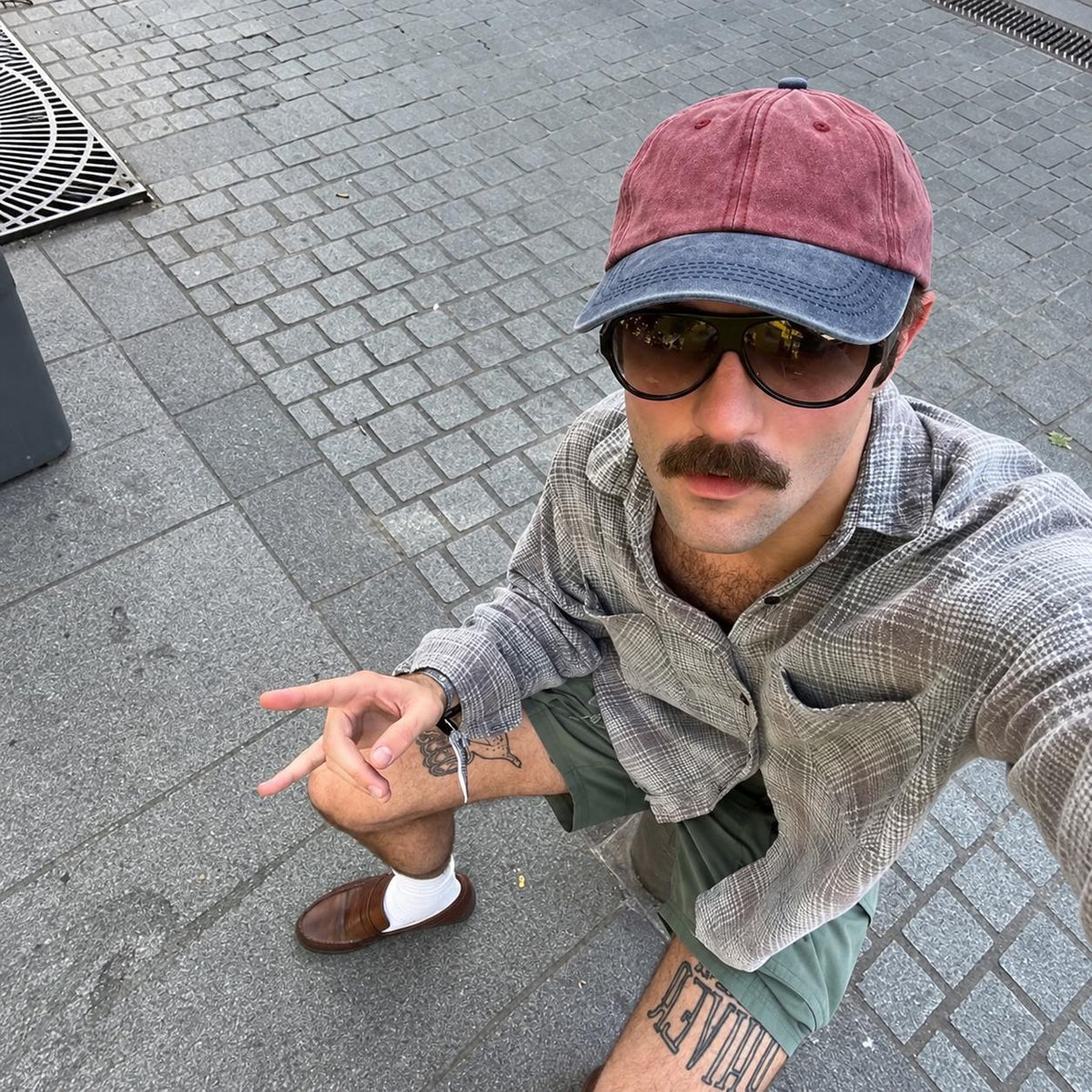 A man wearing a dusty rose and blue baseball cap, dark sunglasses, a gray plaid button-down shirt, and green shorts, taking a selfie on a gray paved street.