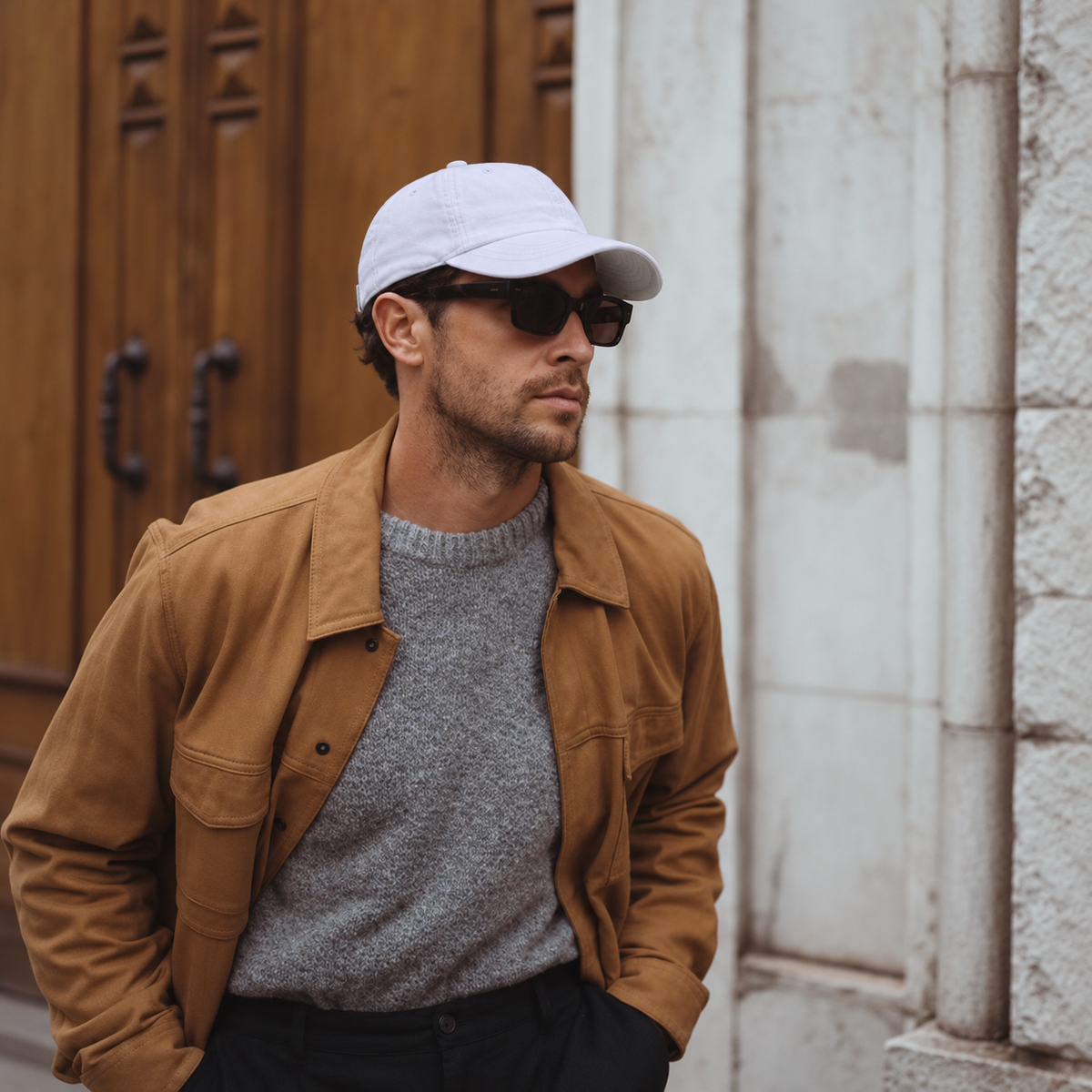 A man wearing a white baseball cap, dark sunglasses, a brown jacket, and a gray knit sweater, standing outdoors next to a wooden door and a stone wall.