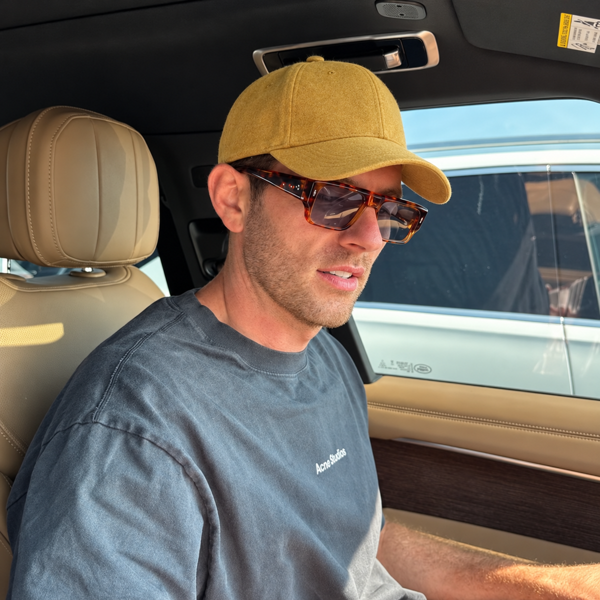 A man wearing a mustard yellow baseball cap, brown tortoiseshell sunglasses, and a gray t-shirt sitting inside a car with beige leather seats.
