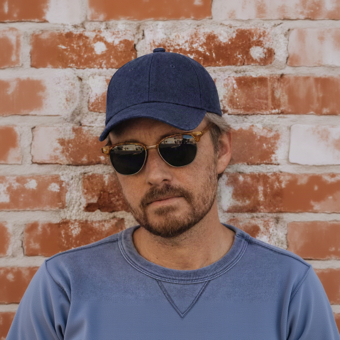 A man wearing a navy blue baseball cap, gold-framed sunglasses with dark lenses, and a blue-gray crewneck sweatshirt, standing against a rustic red-brown brick wall.
