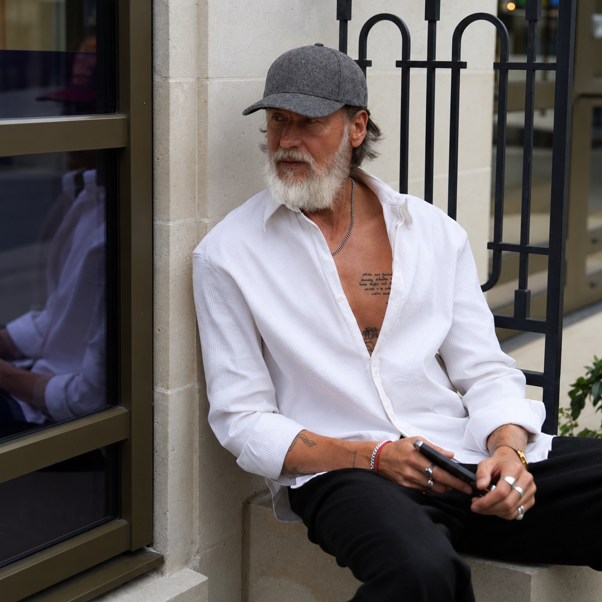A man with a white beard and gray cap sits on a stone ledge wearing an open white button-down shirt, black pants, and holding a black smartphone. He is posed against a beige stone wall with a black metal gate in the background.
