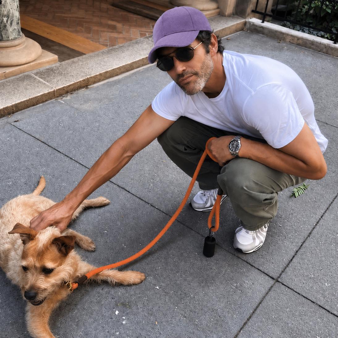 A man wearing a purple baseball cap, white t-shirt, and olive green cargo pants crouches on a stone sidewalk, petting a small tan dog on an orange leash.