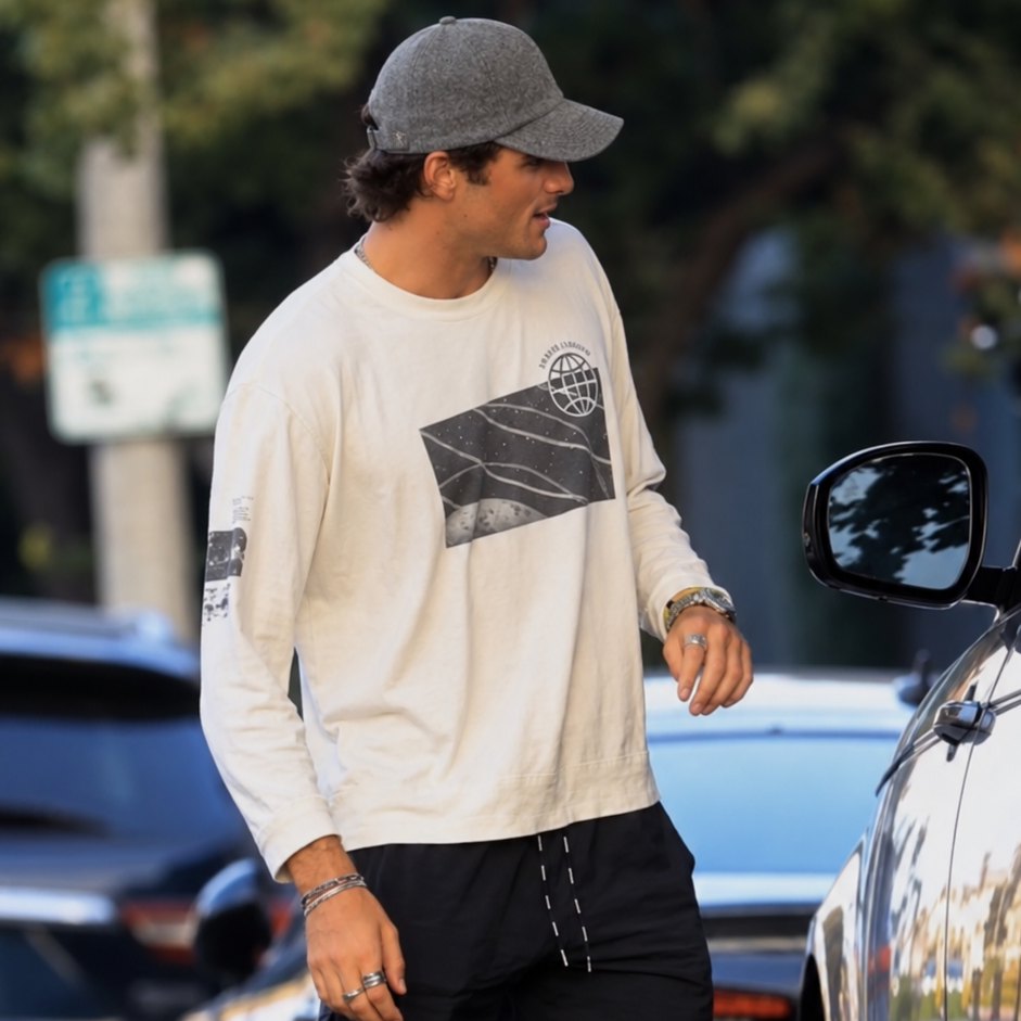 A man walking outdoors on a street wearing a cream long-sleeve graphic shirt, black shorts, a gray baseball cap, and blue sneakers.